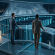 Deux dirigeants d'entreprise dans une salle de serveurs futuriste surplombant une infrastructure d'IA illuminée depuis une mezzanine vitrée, symbolisant la gouvernance et la supervision de l'IA.