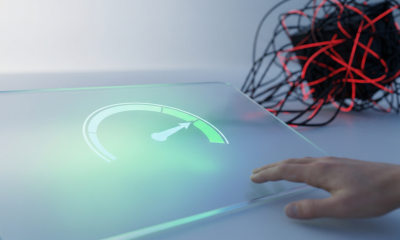 A conceptual photograph showing a hand approaching a transparent glass interface with a glowing green speedometer gauge and an upward-pointing arrow. The background shows an out-of-focus, tangled knot of black wires and red light.