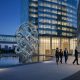 Business professionals shaking hands in a modern glass-walled corporate plaza next to a large glowing geometric sculpture representing AI connectivity and consolidation.