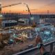 A widescreen, photorealistic photograph captures a construction supervisor overlooking a massive data center project at dusk. From an elevated perspective, the supervisor holds a transparent, glowing digital tablet displaying a holographic 3D blue and orange schematic with floating data nodes. Below, cranes, heavy machinery, steel frameworks, cooling towers, and electric substations are active under a cinematic sunset sky, symbolizing the integration of physical construction and advanced AI orchestration.