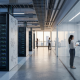 A photorealistic visualization of modern enterprise AI infrastructure. A large corridor features floor-to-ceiling server racks glowing with subtle blue lights on the left. On the right, beyond glass partitions, a corporate strategy team of three (wearing business casual attire) collaborates. One person gestures toward a large, interactive wall display showing a network of abstract nodes and data streams (representing strategic scaling phases like PILOT and CAPABILITY), while another holds a tablet. The background looks out onto a city skyline at dusk.