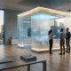 A widescreen, photorealistic image of a modern, glass-walled financial office at sunset, where three professionals stand before a glowing digital display analyzing complex data visualizations and charts.