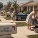 AI-generated image (GPT-2): A 1960s suburban street where identical Stepford-style wives clean cars in repeating rows, with a ‘3081 Stepford St’ mailbox in the foreground.