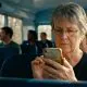 A close-up, photorealistic shot of an older woman with short grey hair and glasses intensely focused on her smartphone while sitting on a public bus. Other passengers are visible in the soft-focus background, emphasizing a shared, everyday commuting environment.