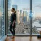A photorealistic widescreen image of a woman standing in a modern high-rise apartment overlooking a city at sunrise, with holographic AI neural networks and data visualizations floating in the foreground.