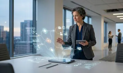 A professional woman in a modern office environment interacting with a holographic interface displaying data nodes and collaborative icons above a tablet.