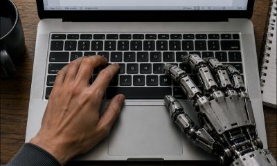 AI-generated image (GPT-2): Overhead view of a laptop on a wooden desk, with a human left hand and an industrial robotic right hand positioned on the keyboard, a text document open on the screen, and a notebook, pen, and coffee mug visible beside the computer.