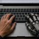 AI-generated image (GPT-2): Overhead view of a laptop on a wooden desk, with a human left hand and an industrial robotic right hand positioned on the keyboard, a text document open on the screen, and a notebook, pen, and coffee mug visible beside the computer.