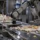 AI-generated image (GPT-2): A metal industrial robotic arm presses a flat circular plate into a decorated cake on a stainless steel conveyor belt, crushing it into a spread of frosting and crumbs, while intact cakes move toward it in a factory setting.