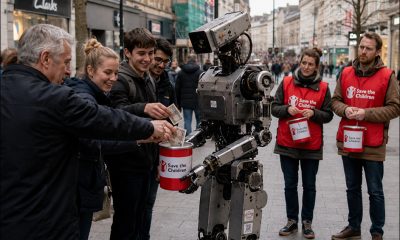 AI-generated image (GPT-2) - An industrial humanoid robot holds a Save the Children donation tin on a British high street as several passers-by place five pound notes into it, while nearby human volunteers in red Save the Children vests stand with their own collection tins and watch with puzzled expressions.