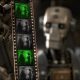 AI-generated image (GPT-2 + Photoshop): an industrial humanoid robot, heavily blurred, holds a vertical strip of 70mm film close to the camera, with the film frames in sharp focus showing a man and a woman standing and conversing. Every third frame is tinted green and the others appear monochrome.