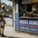AI-generated illustration (GPT-2 + Photoshop): A humanoid industrial robot stands on a sunlit city sidewalk facing a small check-cashing kiosk, while a wolf-like attendant leans from the service window reading a sports magazine as pedestrians and traffic pass in the background.