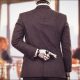 GPT Image 2 (AI-generated image): A humanoid robot wearing a dark suit stands with its back to the camera at a conference table, with one mechanical hand with fingers crossed behind its back. Blurred business attendees sit on both sides, with papers, glasses, windows, and a city view in the background.