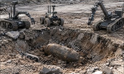 AI-generated image by GPT-Image-2: Three bomb disposal robots of different designs stand around a rubble-lined crater containing a large, partially buried unexploded bomb. The surrounding terrain consists of broken concrete, stones, dirt and damaged pavement, with ruined masonry visible in the background and scattered debris extending to the image edges.