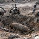 AI-generated image by GPT-Image-2: Three bomb disposal robots of different designs stand around a rubble-lined crater containing a large, partially buried unexploded bomb. The surrounding terrain consists of broken concrete, stones, dirt and damaged pavement, with ruined masonry visible in the background and scattered debris extending to the image edges.