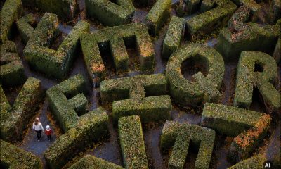 AI-generated image: a woman and young child walk together through a maze of tall green hedges shaped into large, randomly oriented alphabet letters. Narrow pathways separate the hedges, with fallen autumn leaves scattered across the ground. Viewed from above. Image created using GPT Image 2.
