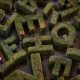 AI-generated image: a woman and young child walk together through a maze of tall green hedges shaped into large, randomly oriented alphabet letters. Narrow pathways separate the hedges, with fallen autumn leaves scattered across the ground. Viewed from above. Image created using GPT Image 2.