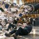 AI-generated image (GPT Image 2): chaotic scene in a university library lined with bookshelves, where a large industrial robotic arm blasts a torrent of books, journals and loose papers through a wide hose. Several adults are knocked off balance or fall to the floor, while others watch from behind desks. Scattered printed materials cover the floor.